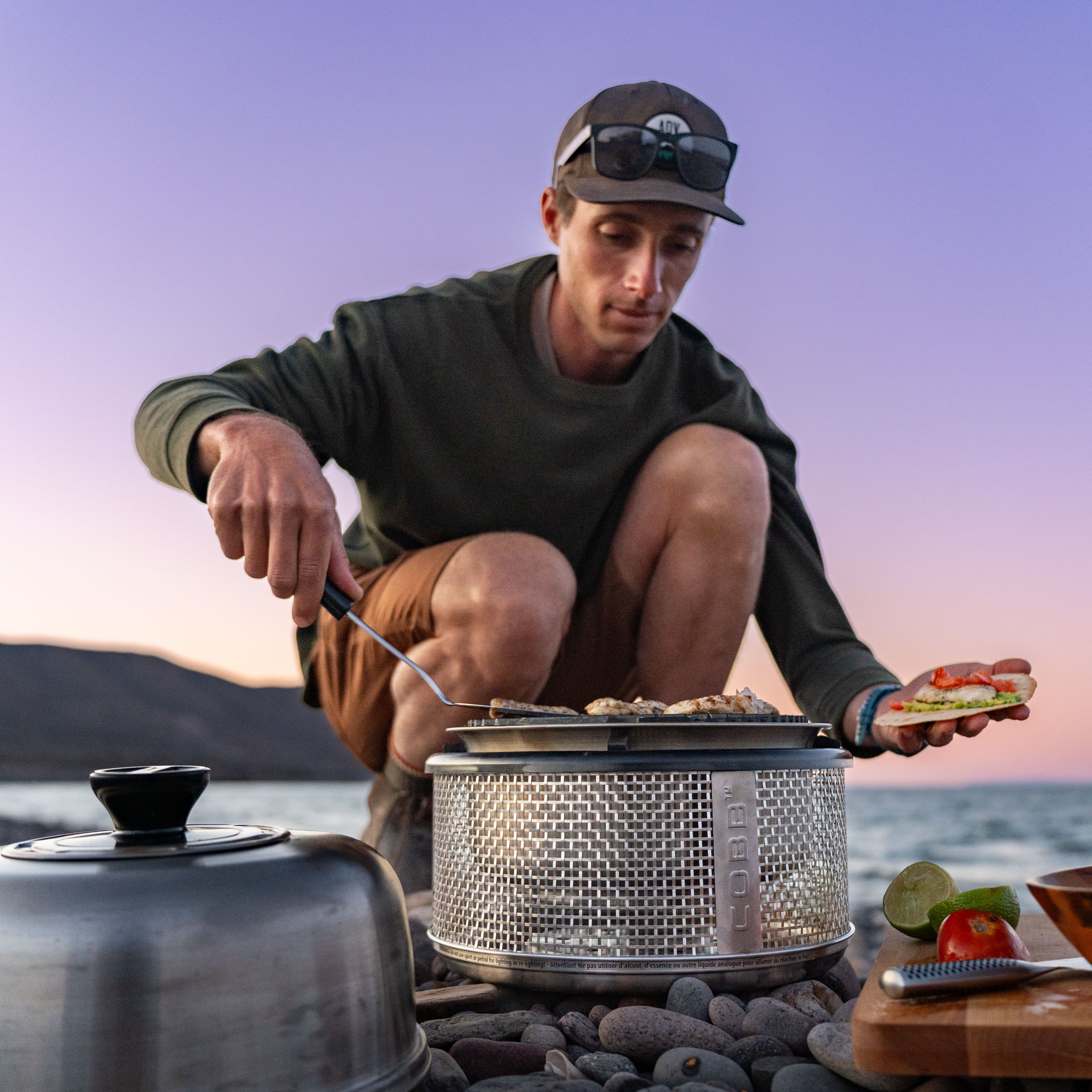 Person cooking outdoors on a rocky beach with a sunset in the background